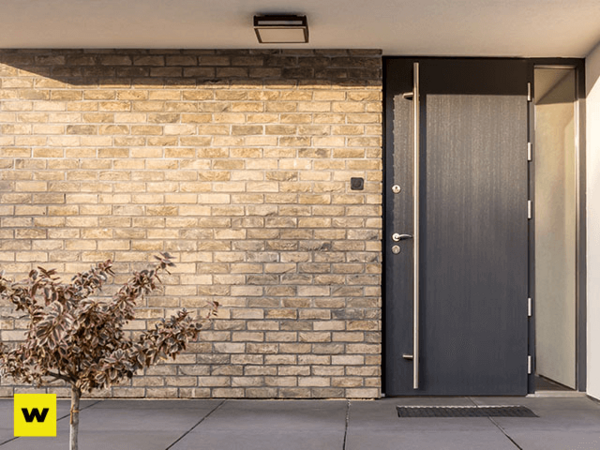 Moderne Haustür aus dunklem Aluminium neben heller Klinkerfassade und kleinem Busch.